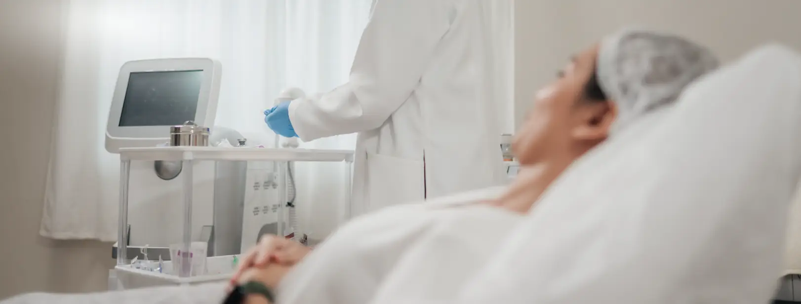Patient reclines on a treatment bed while a clinician stands beside medical equipment in a clinic room.