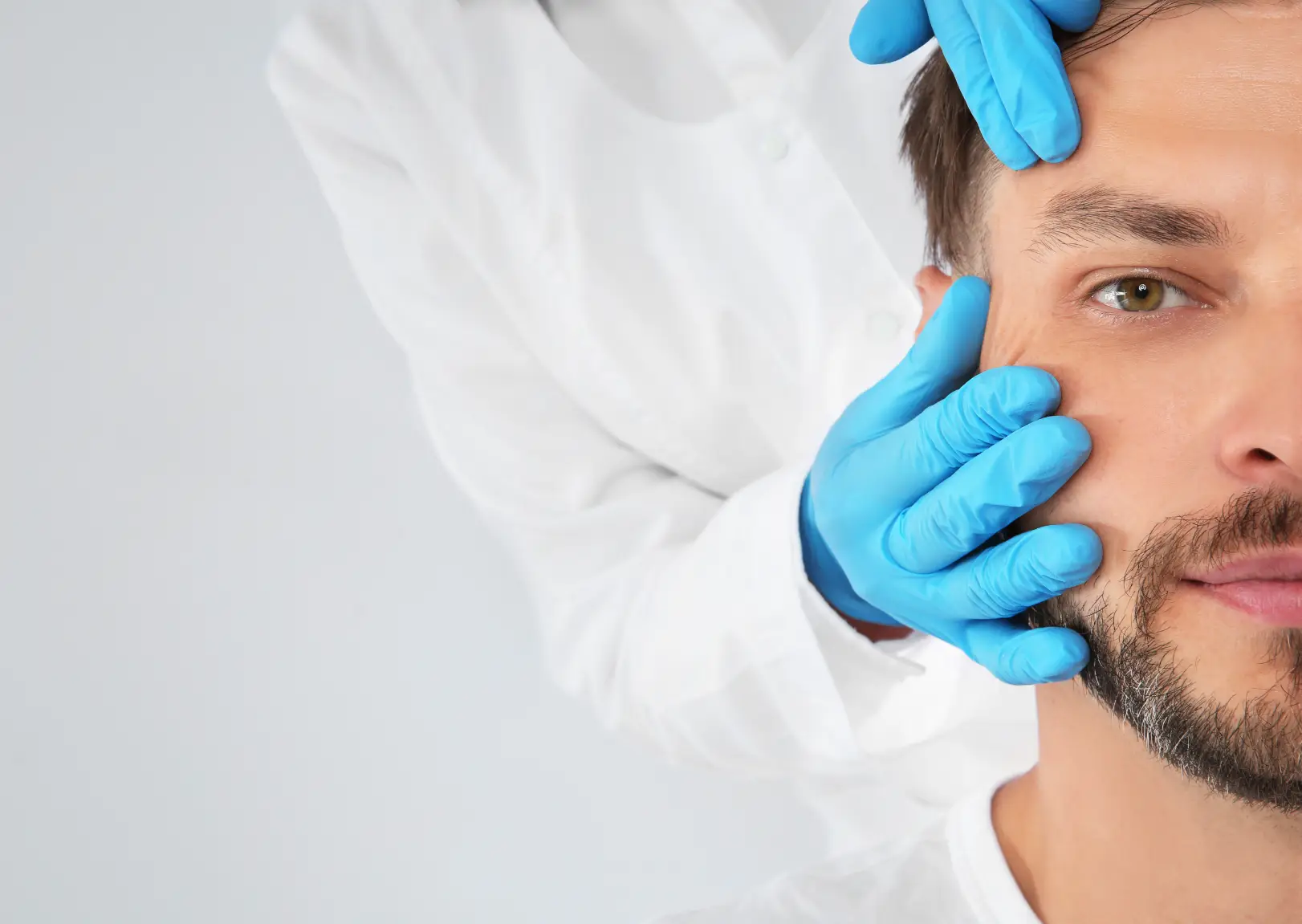 Gloved hands gently position a man's face during a facial examination against a light background.