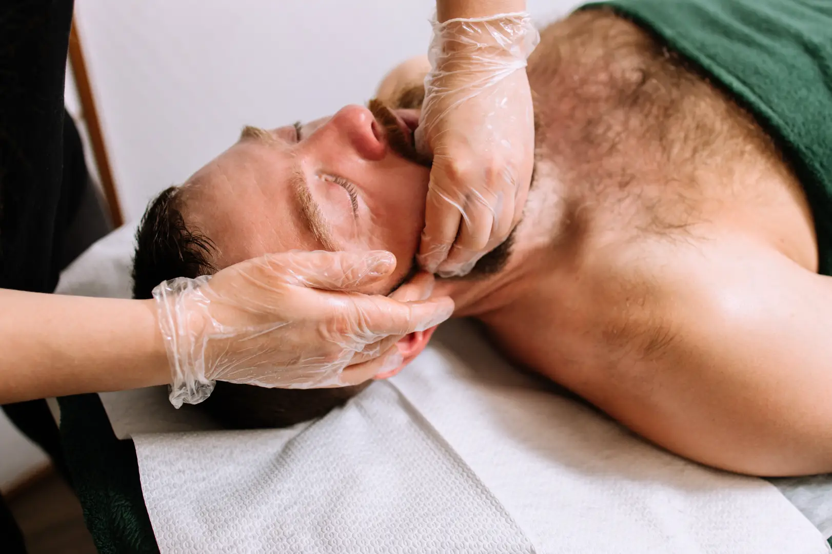 Person lying on a treatment bed while gloved hands apply pressure along the jaw and cheek area.