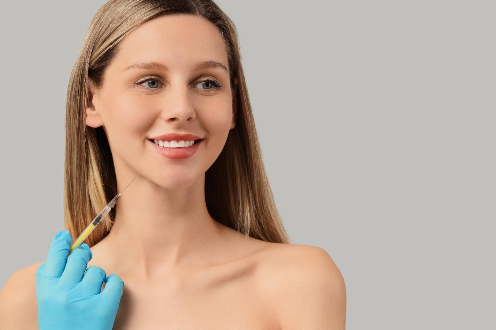 Gloved hand holding a syringe near the jawline of a smiling person facing slightly to the side.